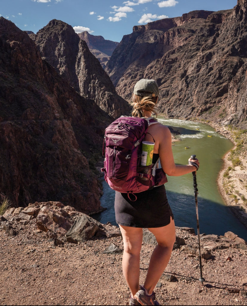 Woman wearing purple backpack stands facing the Colorado River while hiking in the Grand Canyon.