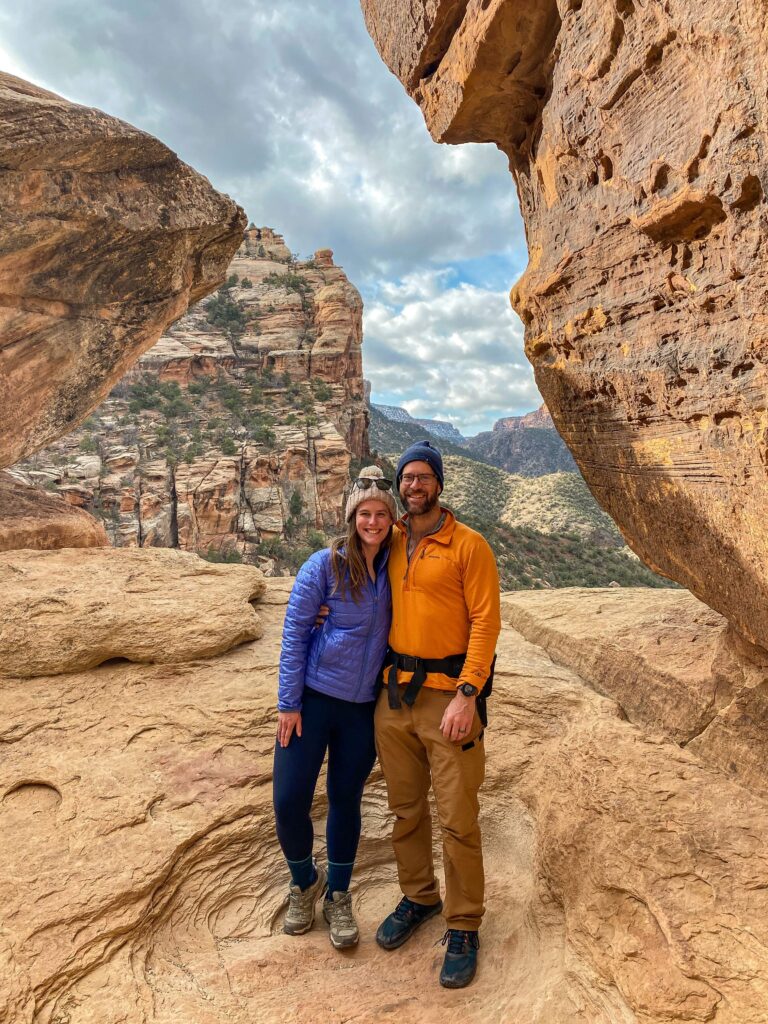 A couple wearing hiking gear including winter hats, a periwinkle jacket, and orange pullover stands on a sandstone rock with a canyon behind them at Devils Kitchen in Colorado National Monument.
