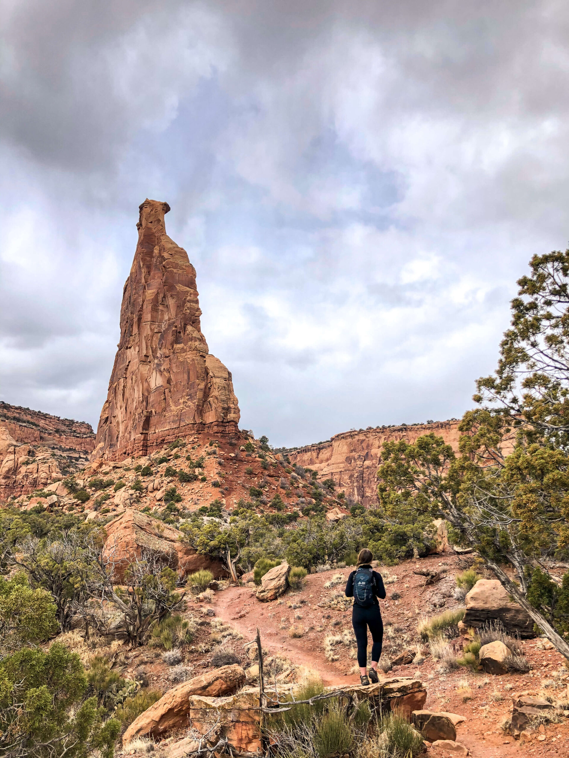 Woman wearing a backpack stands on a rock in front of Independence Monument while hiking in Colorado National Monument.