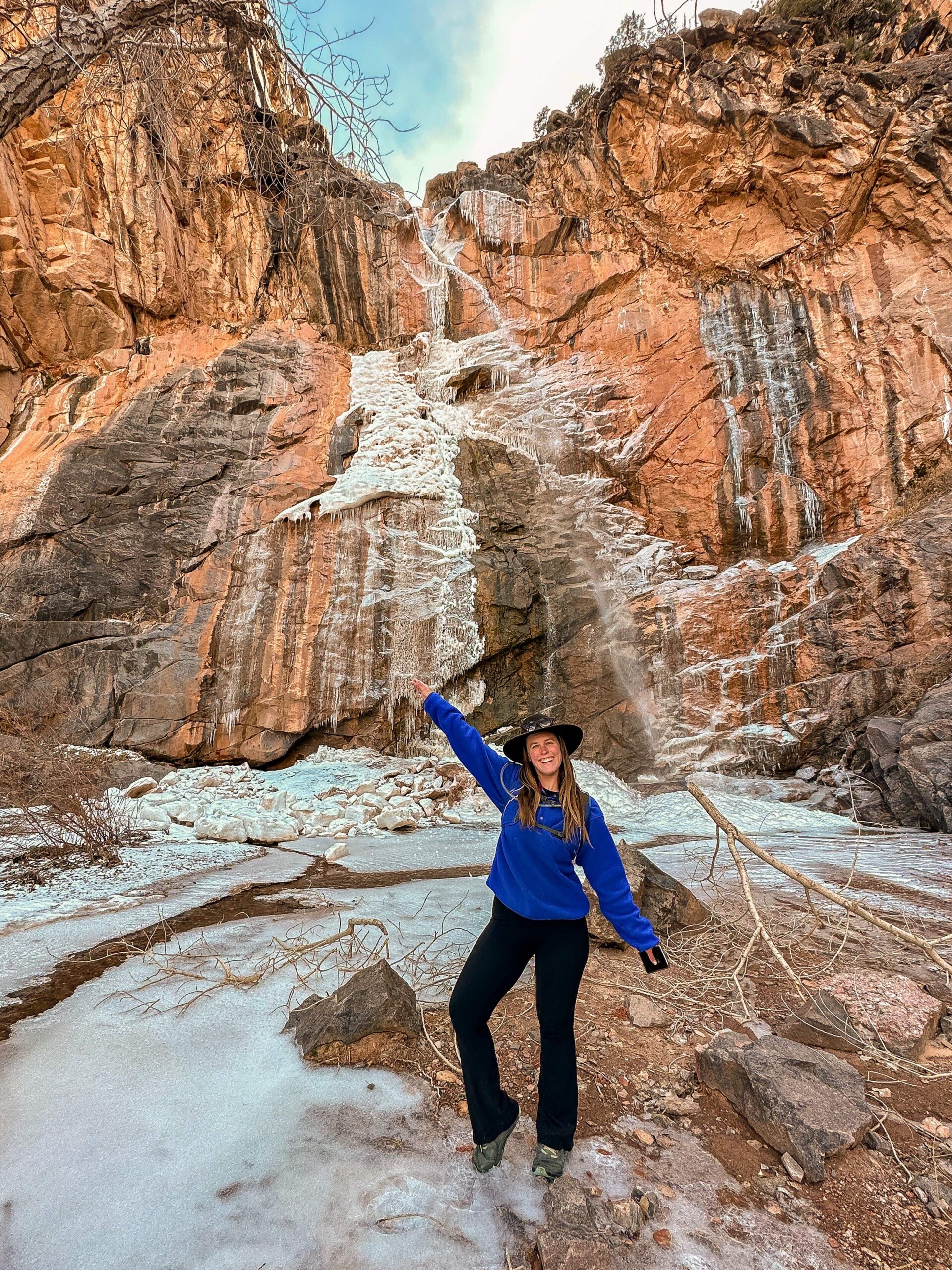 Woman wearing a cobalt blue sweatshirt and yoga pants stands in front of a waterfall in No Thoroughfare Canyon in Colorado National Monument.