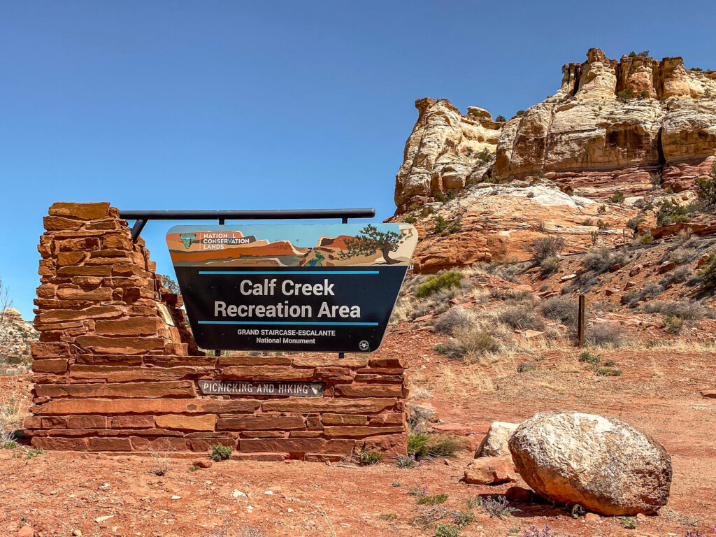 National Conservation Lands sign for Calf Creek Recreation Area with red rocks in the background.