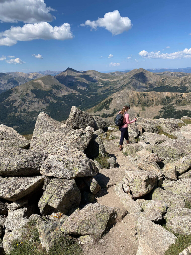 Female hiker wearing a pink top and hiking poles walks past large granite boulders with mountains in the distance in Colorado.
