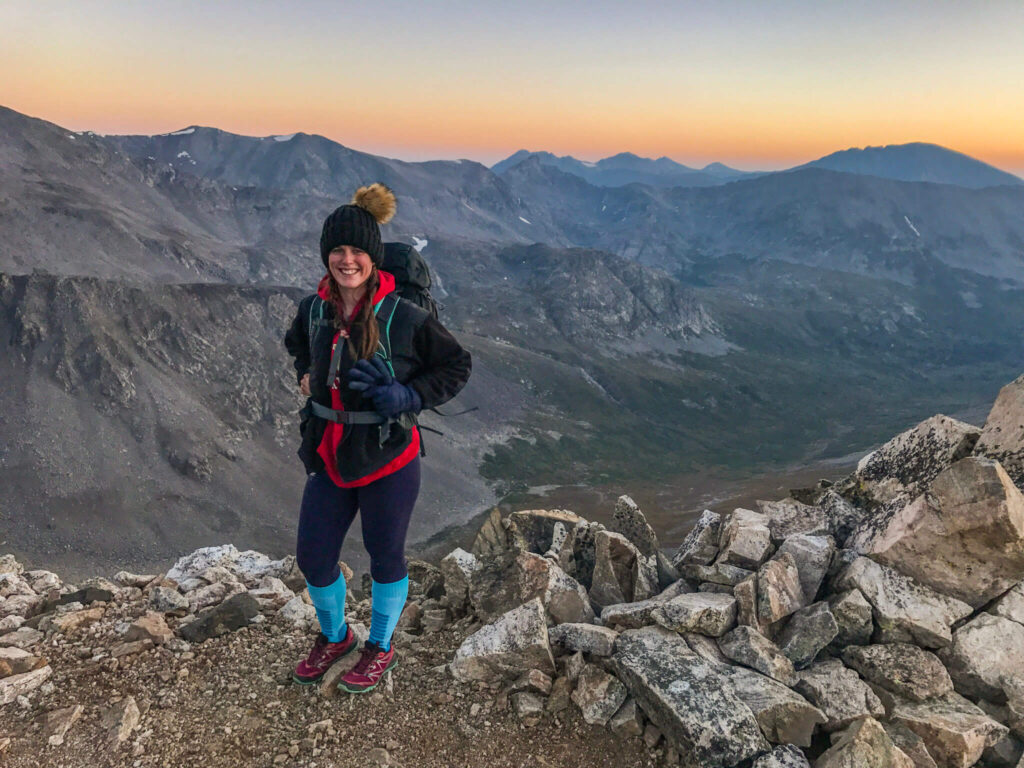 Woman wearing a ridiculous puffy hat, sweatshirt, and north face jacket with leggings and tall blue socks climbing a 14'er in Colorado at sunrise.