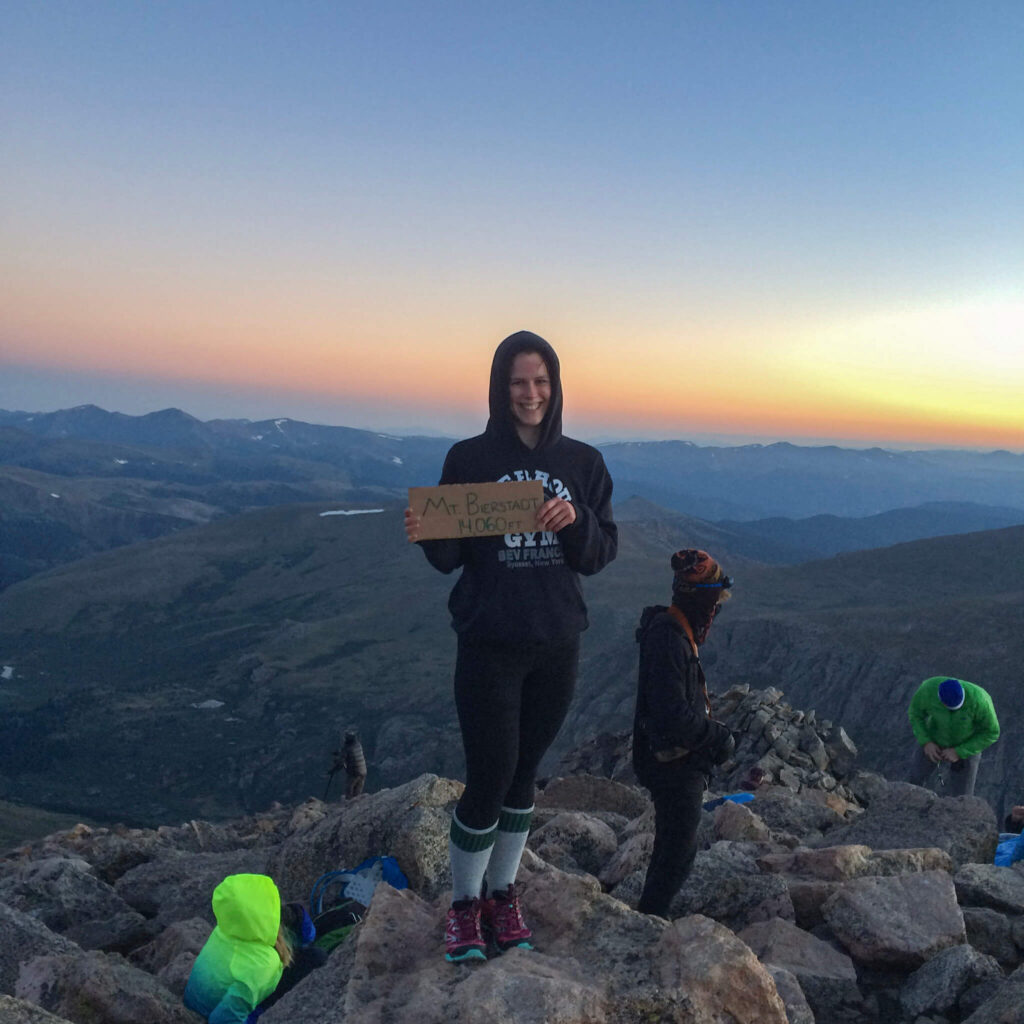 Woman wearing a black sweatshirt with the hood up stands on top of Mt. Bierstadt at sunrise while climbing her first Colorado 14'er.