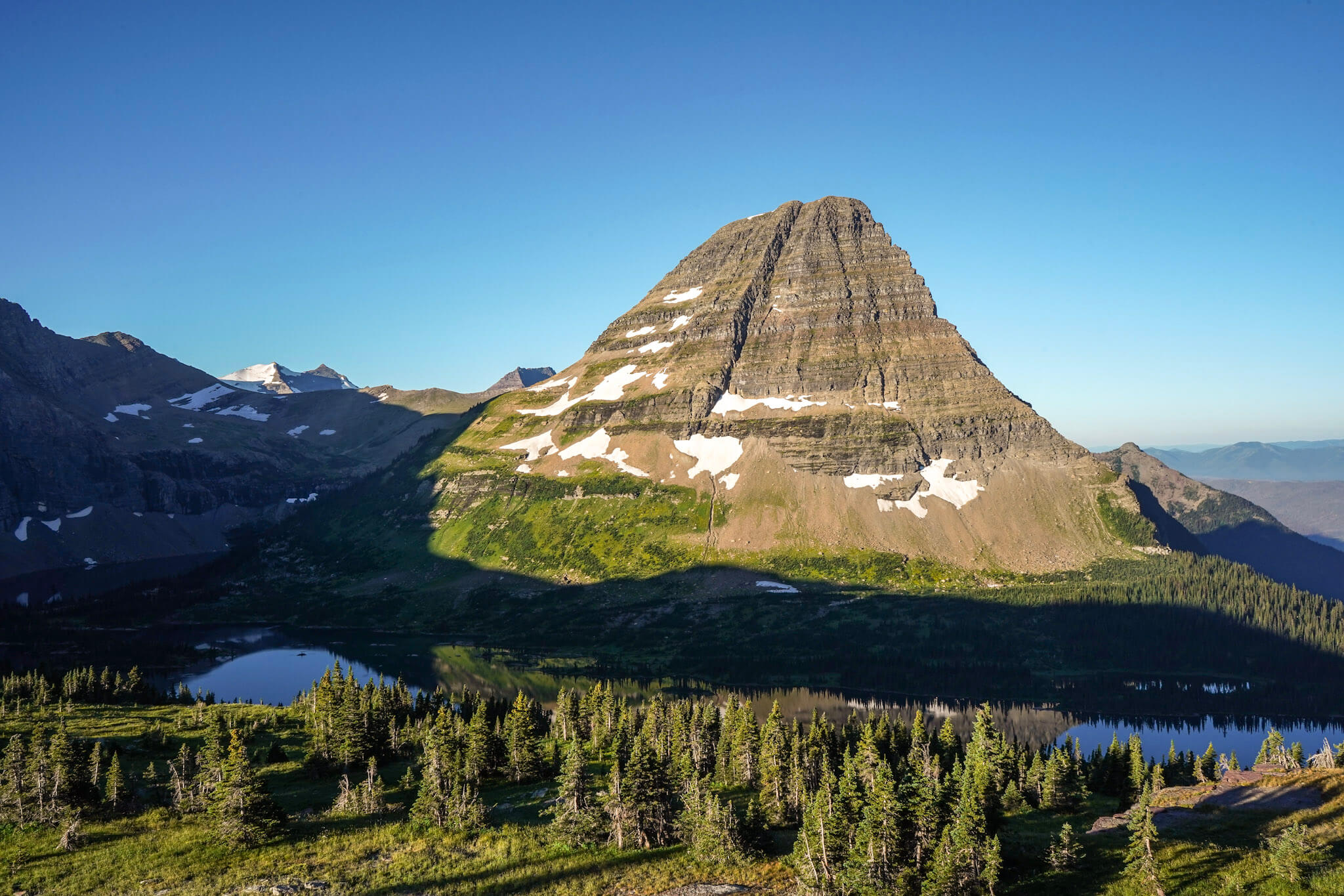 A lone mountain with patches of snow towers over Hidden Lake in Glacier National Park surrounded by trees along the shore with a bright blue sky.