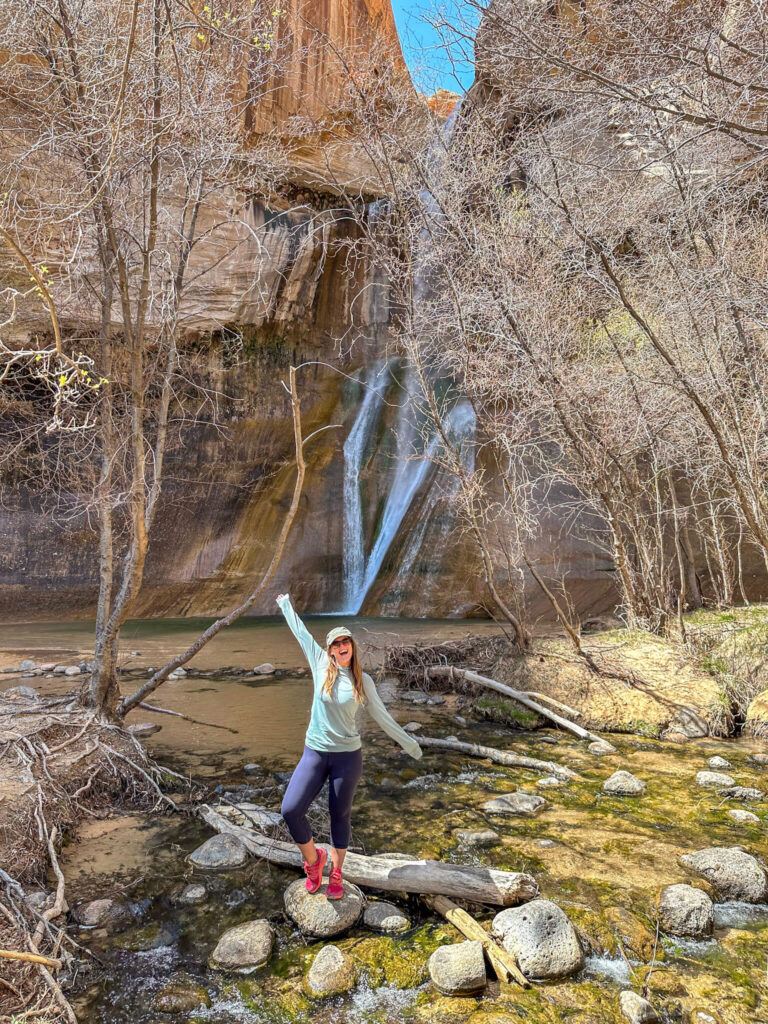 Woman wearing hot pink Altra trai runners in front of bare cottonwood trees and a desert canyon waterfall.