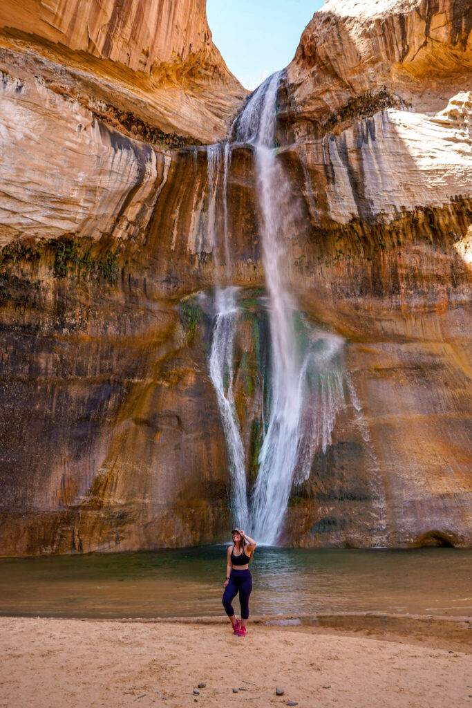 Woman wearing a sports bra, leggings, and hot pink trail runners stands with her hand on the brim of a baseball cap in front of Lower Calf Creek Falls in Grand Staircase-Escalante National Monument.