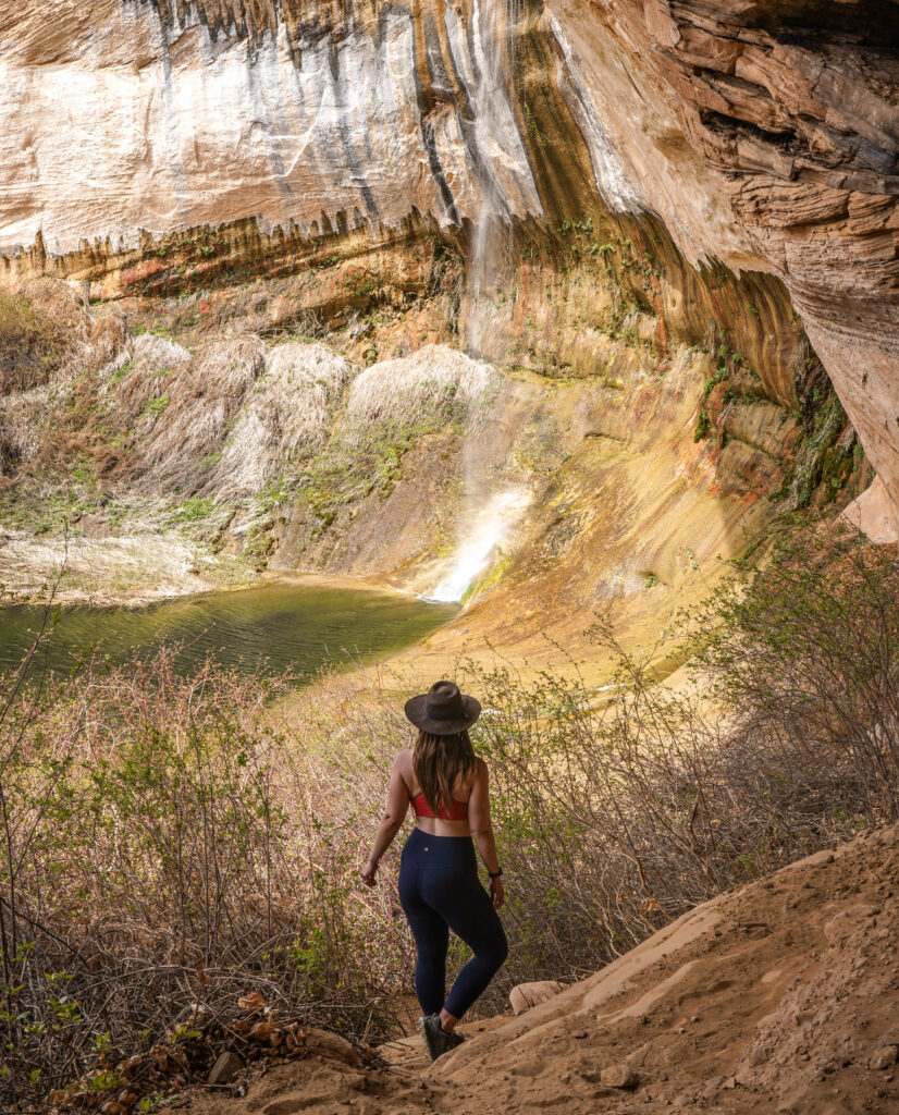 Woman wearing a wide-brim black hat, orange sports bra, and navy blue Lululemon leggings stands with her knee popped in front of Upper Calf Creek Falls in Grand Staircase-Escalante National Monument. The waterfall is fairly small and splashes down a sandstone wall to a green pool.