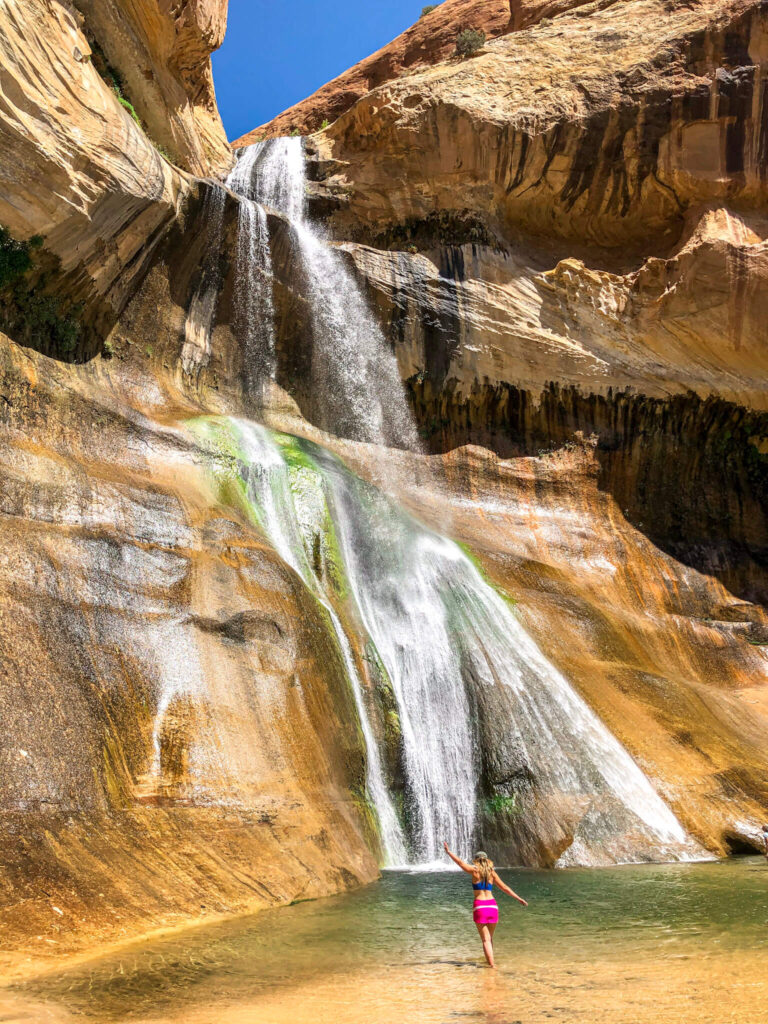 Woman wearing a blue sports bra and hot pink skort walking through a pool of water towards Lower Calf Creek Falls in Grand Staircase-Escalante National Monument with one arm over her head and one arm sticking out.