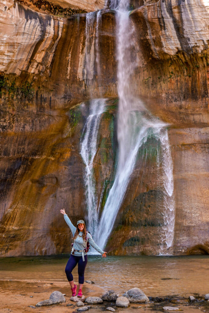 Woman wearing a light blue sun shirt and navy blue leggings with an olive green hat and hot pink trail runners stands with her arms open on a rock in front of Lower Calf Creek Falls in Grand Staircase-Escalante National Monument.