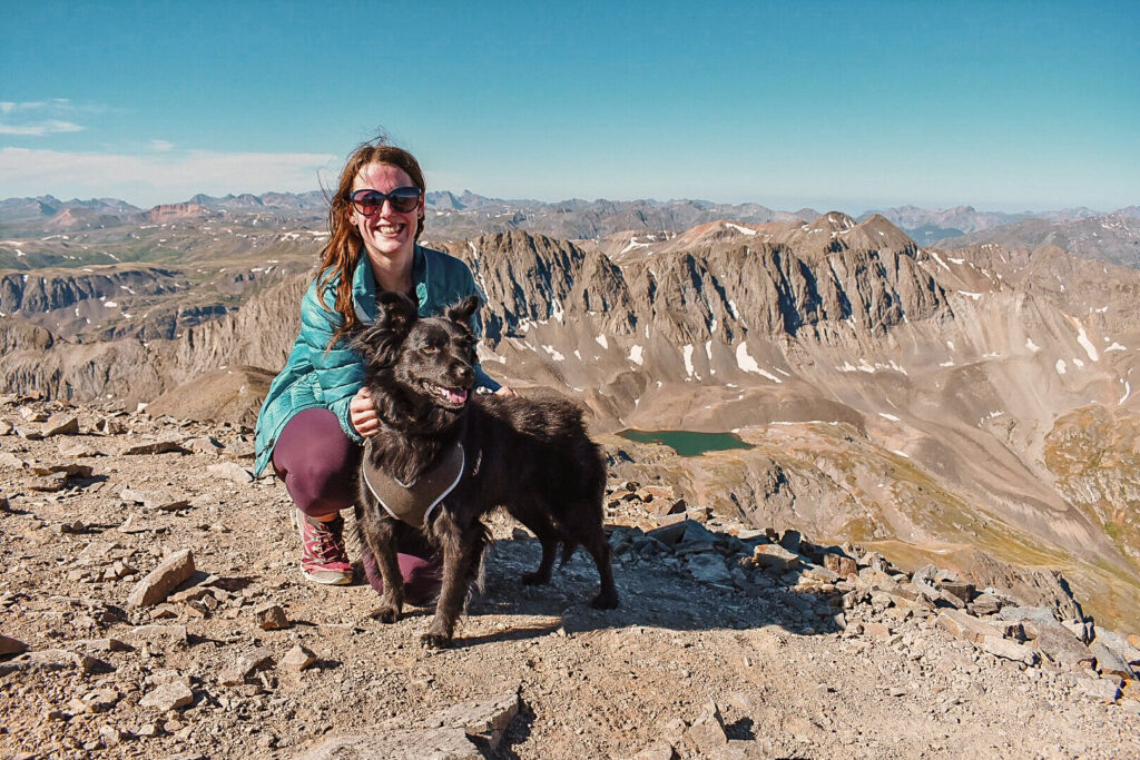 Woman wearing a blue jacket and a black dog stand on top of Handies Peak in the San Juan mountains of Colorado after climbing the dog's first Colorado 14'er.
