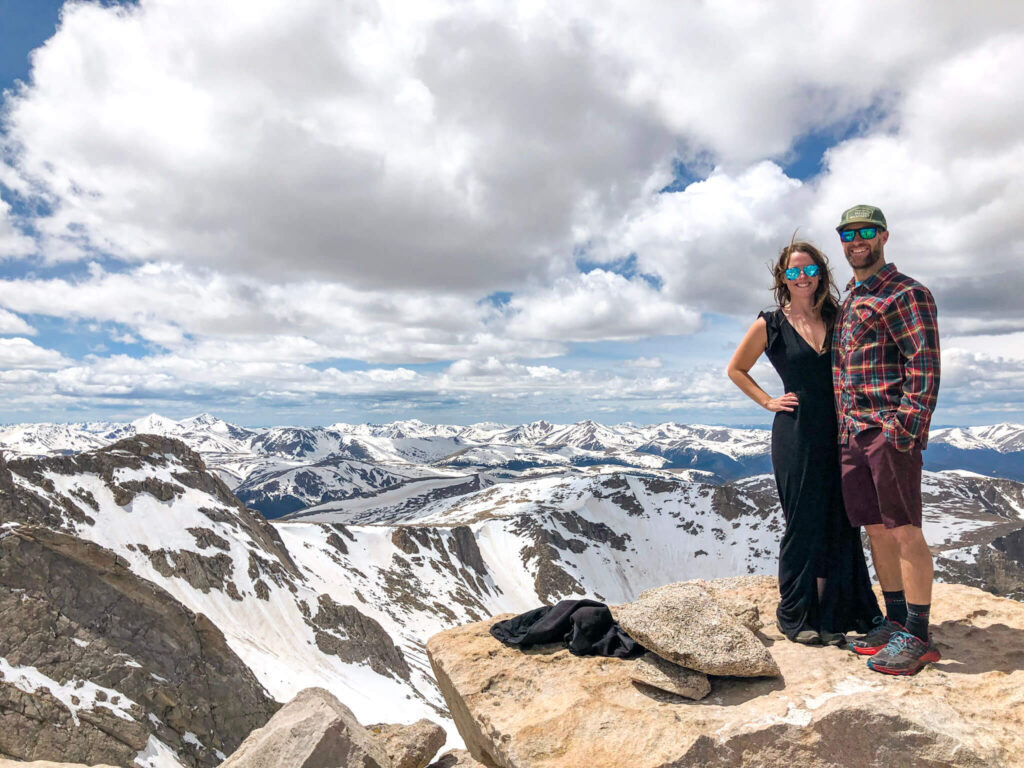A woman wearing a long black dress and sunglasses and a man wearing a flannel shirt and maroon shorts stands on top of a mountain in Colorado with snowcapped mountains behind them.