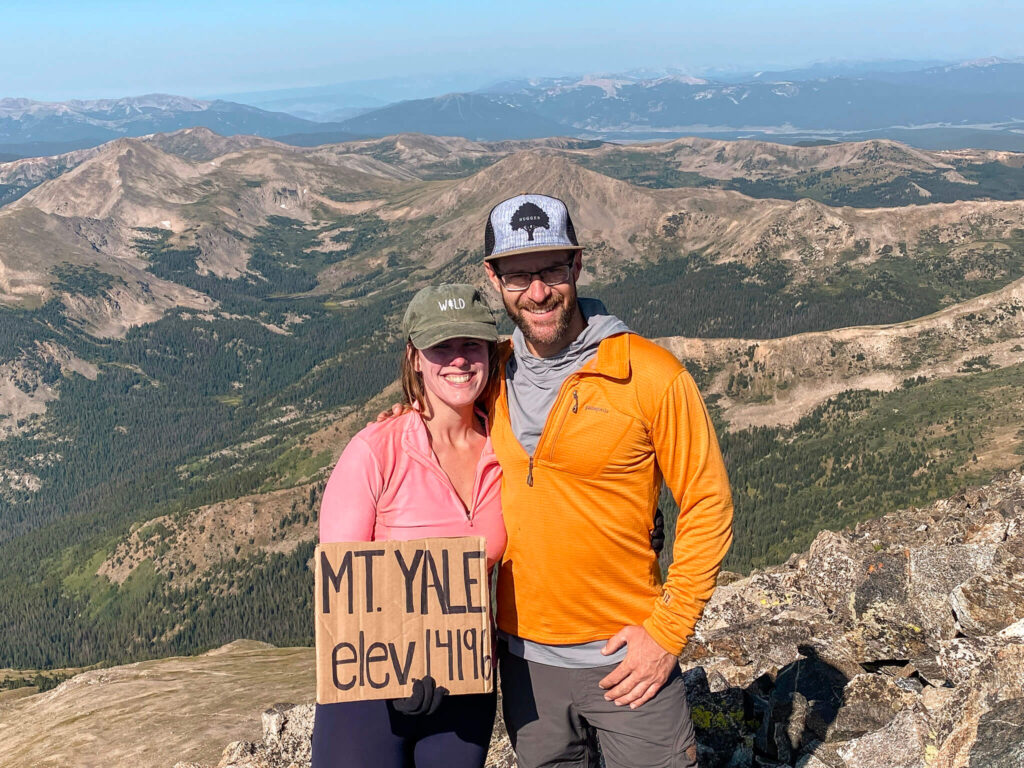 Woman wearing a pink athletic long sleeve top and green hat that says "wild" and man wearing a bright orange Patagonia top stand on top of a 14'er in Colorado with mountains behind the, holding a cardboard sign that says "Mt. Yale elev. 14196."