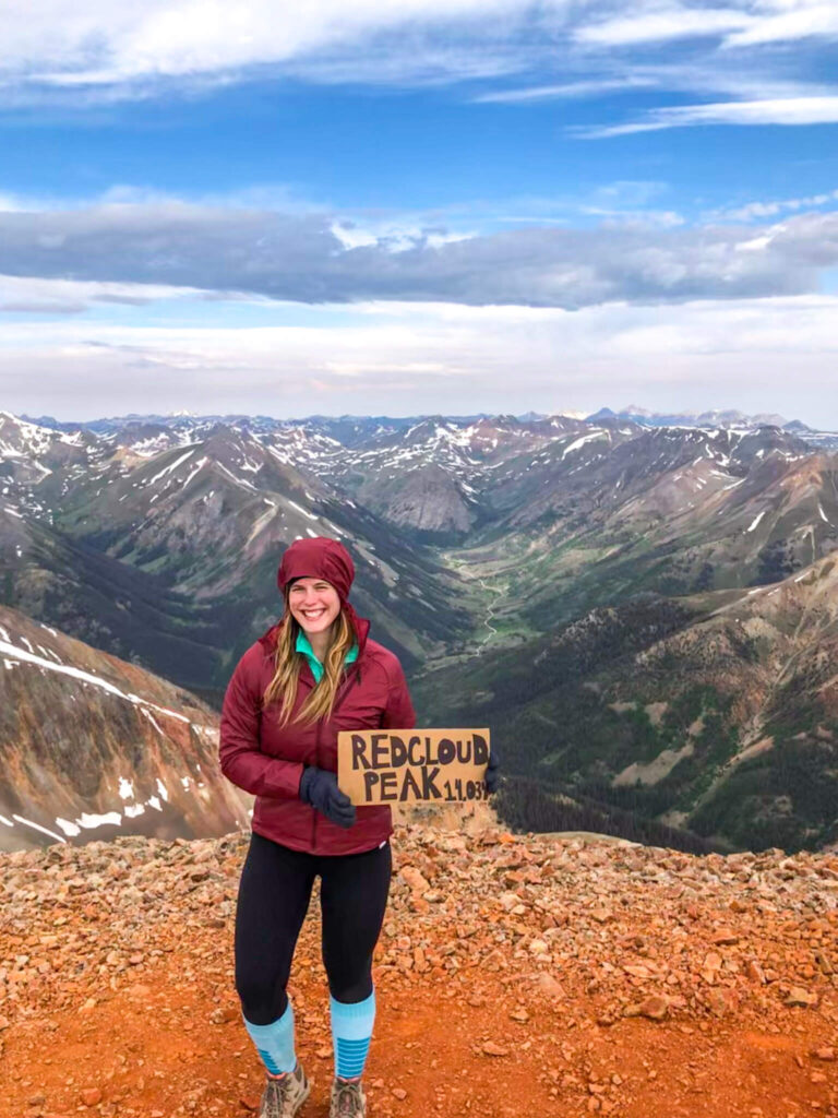 Woman wearing a maroon windbreaker with the hood on stands on top of a red mountain with other mountains in the background holding a cardboard sign that says "Redcloud Peak."