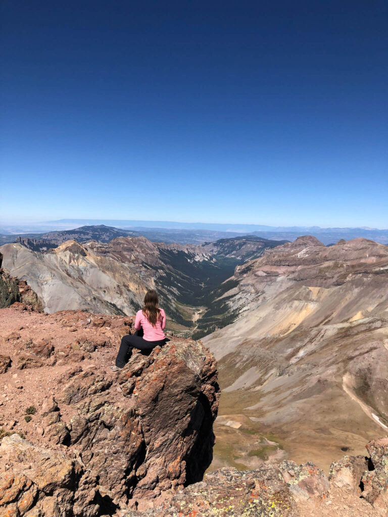 Woman wearing a pink long-sleeve top sits on a rock on Uncompahgre Peak overlooking the San Juan mountains in Colorado.