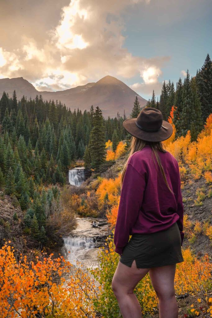 Woman wearing a maroon sweatshirt, mini skirt, and a wide brim black fat stands in front of a waterfall and grove of golden orange aspen trees near Crested Butte in the fall in Colorado.
