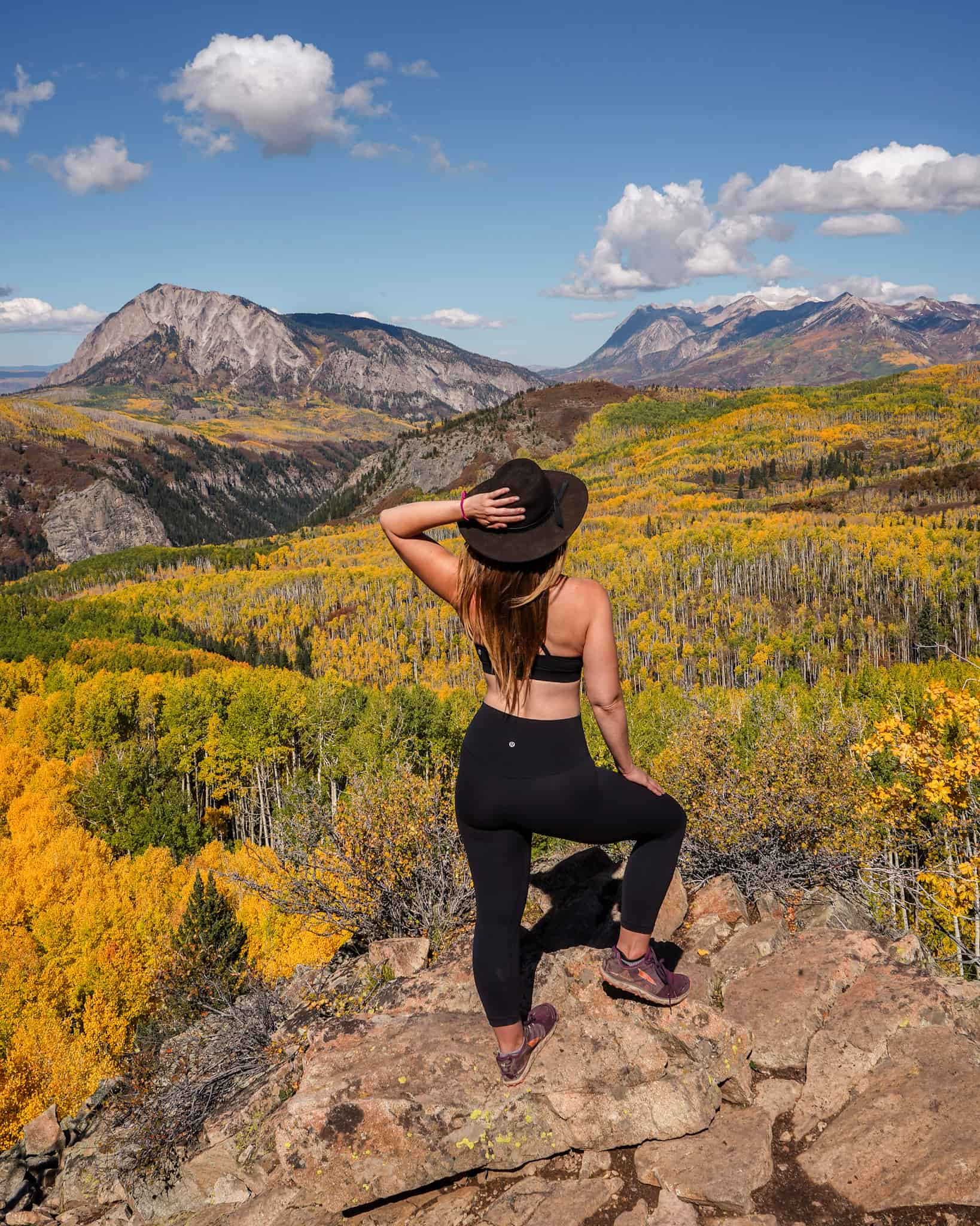 Woman wearing a wide brim black hat and leggings stands with her leg popped on a rock with her hand on her hat overlooking a golden grove of aspens and grey mountains in Crested Butte in the fall in Colorado.