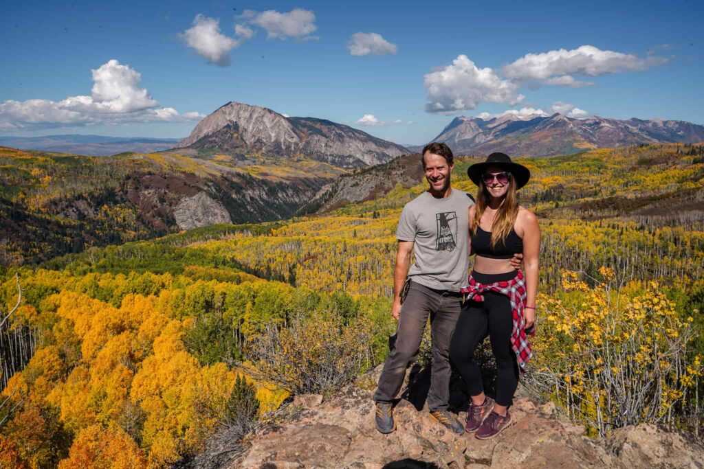 A man in a grey shirt and slacks and a woman with a black sports bra, leggings, and red flannel tried around her waist stand on a rock overlooking an expansive grove of golden aspen trees with jagged mountains in the distance in Crested Butte in the fall.