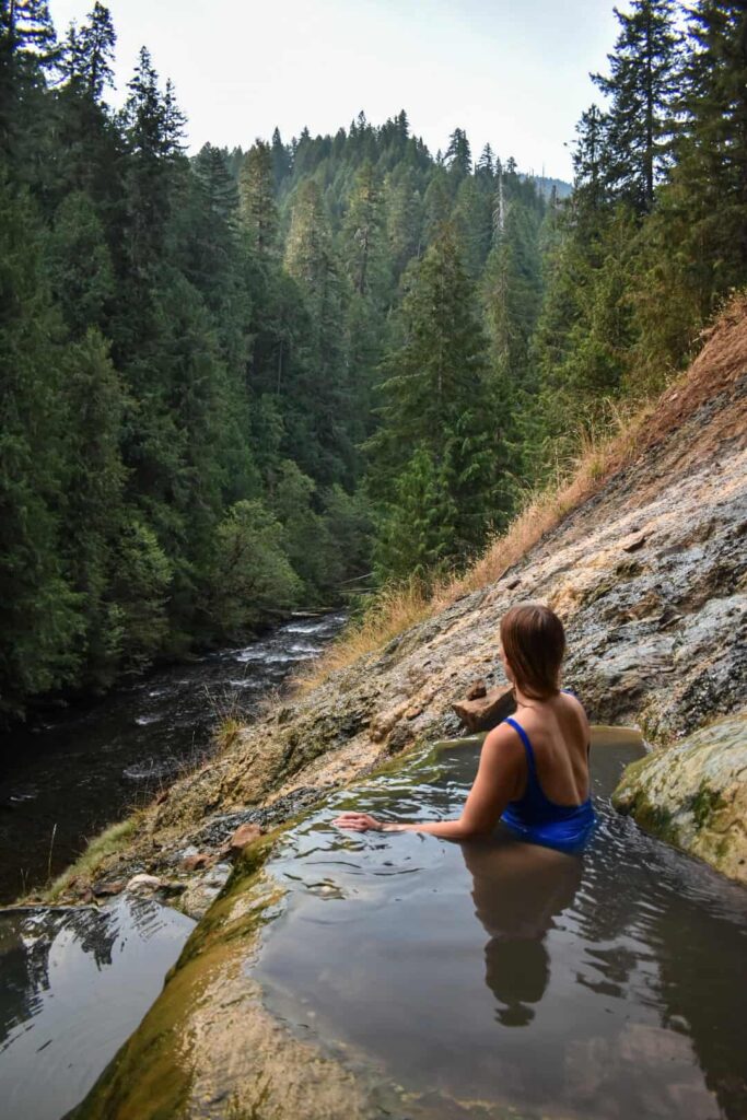 Woman wearing a blue bathing suit sits in a hot spring with a river rushing below.