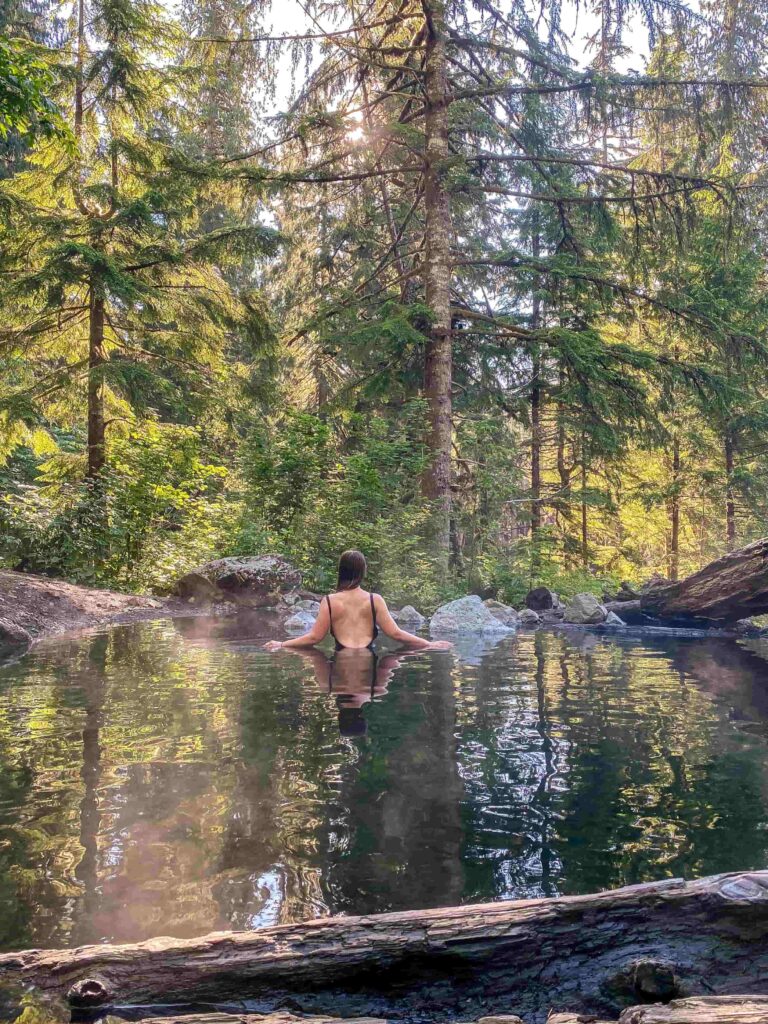 Woman in a black bathing suit sits in a large hot springs pool in a forest with her arms out to her side.