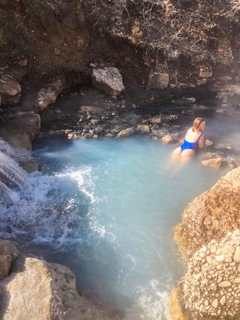 Woman in blue bathing suit leans on the edge of a rock in a blue hot spring with a waterfall.