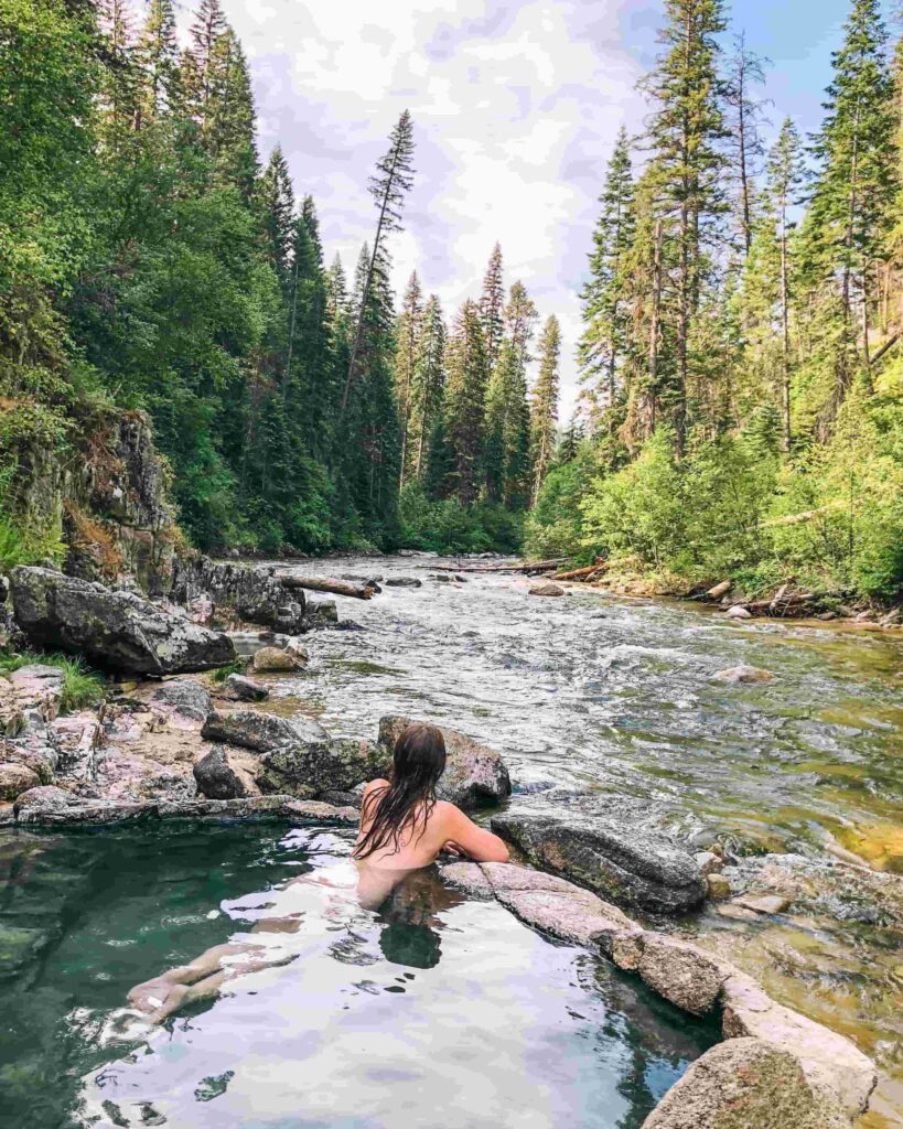 A woman lays in a primitive hot springs overlooking a river surrounded by evergreen trees.