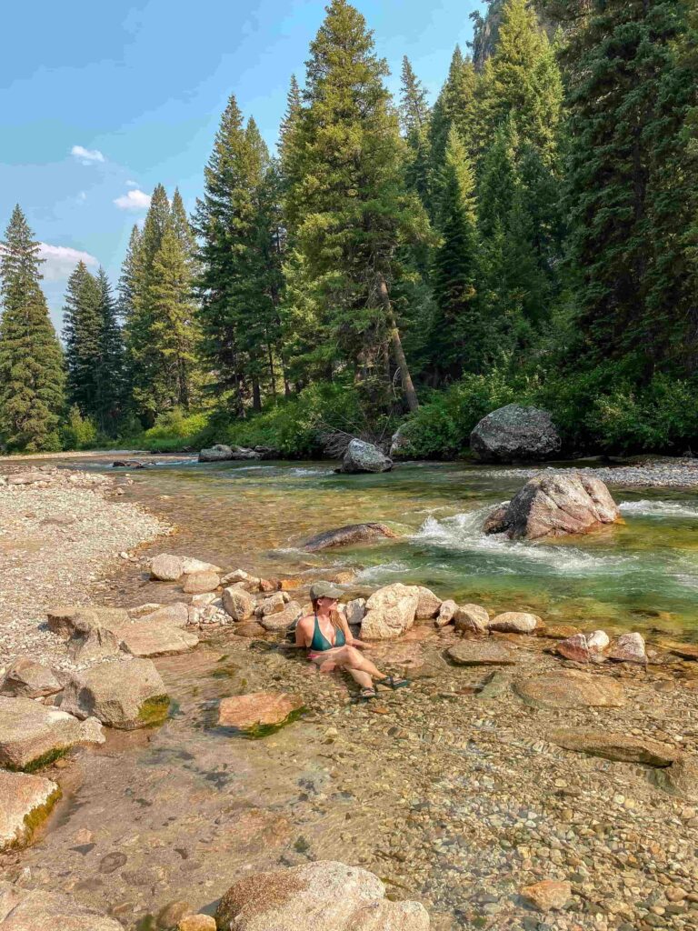 Woman wearing a hat sits in a hot springs next to a river in Idaho surrounded by rocks.