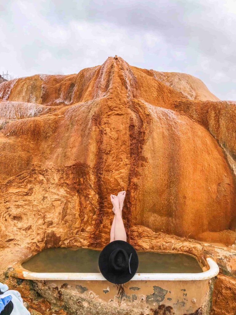 Woman lays in a bath tub that has been overtaken by mineral rich travertine with a black hat and her legs up on the walls of the hot springs.