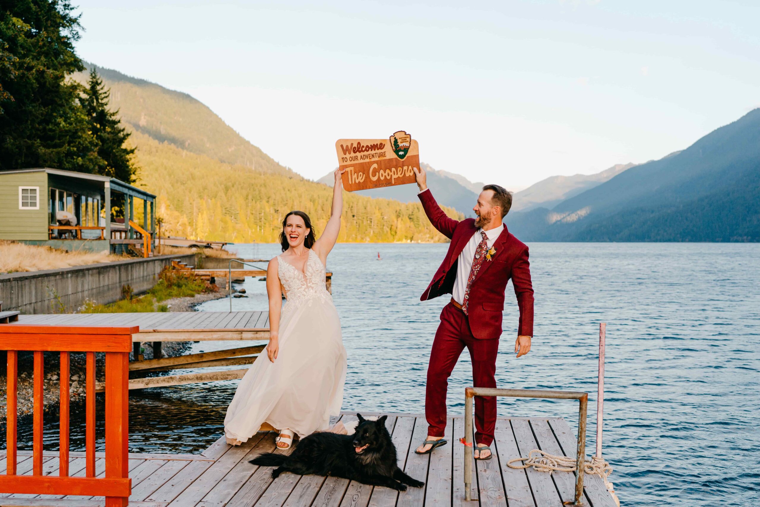 Bride in v-neck chiffon dress and groom in maroon suit hold a sign that says "Welcome to Our Adventure" shaped like a national park sign with a black dog lying in front of them on a dock on the shore of Lake Crescent with Mount Storm King behind them.