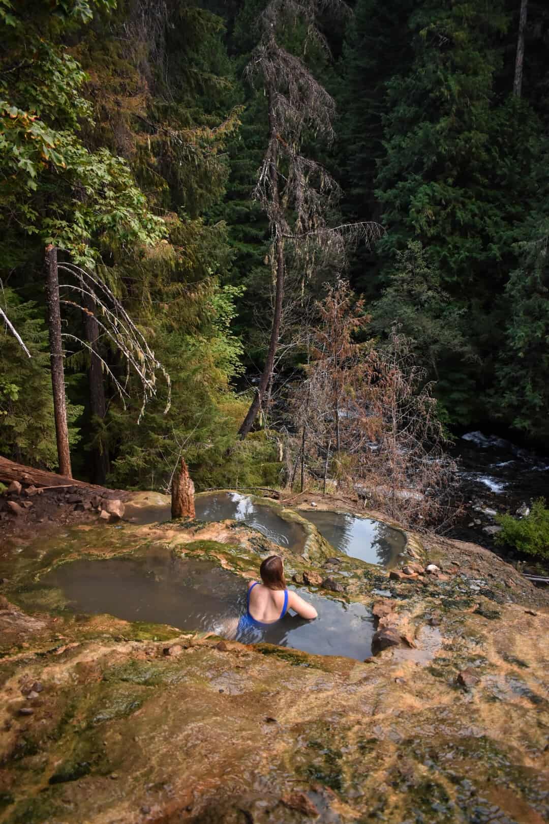 Woman wearing a blue bathing suit sits in a hot spring. It is the top of 3 travertine style hot springs in a forest with a river rushing below.