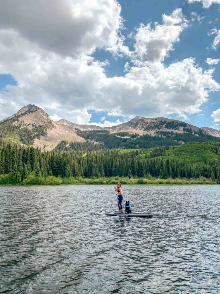 A woman and dog paddleboard on a lake in front of a green forest and mountains in Crested Butte, Colorado.