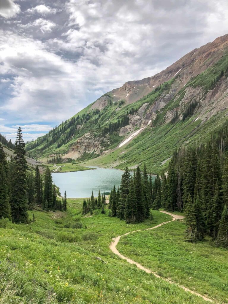 A mountain lake surrounded by evergreen trees with a trail winding towards the lake and granite mountains behind in Crested Butte, Colorado.