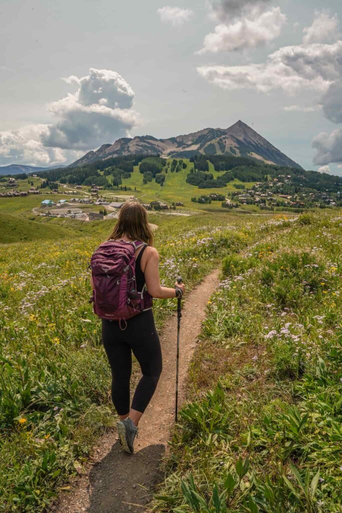 A woman wearing a purple backpack and leggings stands with hiking poles on a trail surrounded by wildflowers with Mt. Crested Butte in the distance.