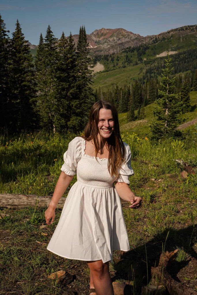 A smiling woman in a white dress with puffed sleeves and smocking stands in front of a green meadow with evergreen trees and mountains behind her in Crested Butte, Colorado.