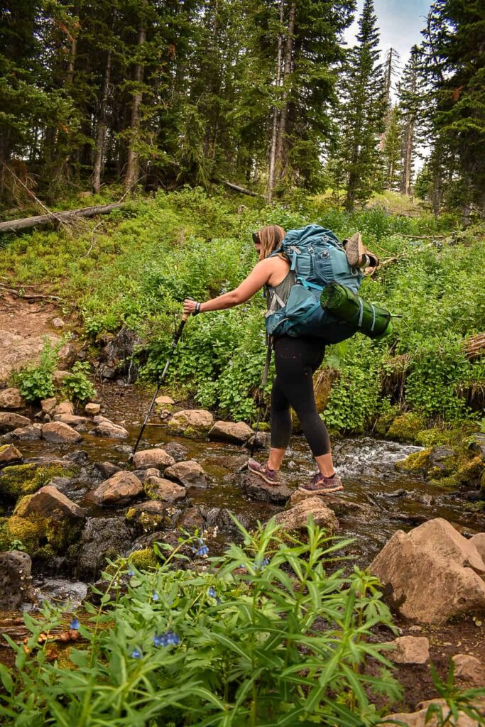 Woman carrying a large blue backpacking pack on an overnight backpacking trip hikes with trekking poles over rocks in a stream through a green forest in Colorado.