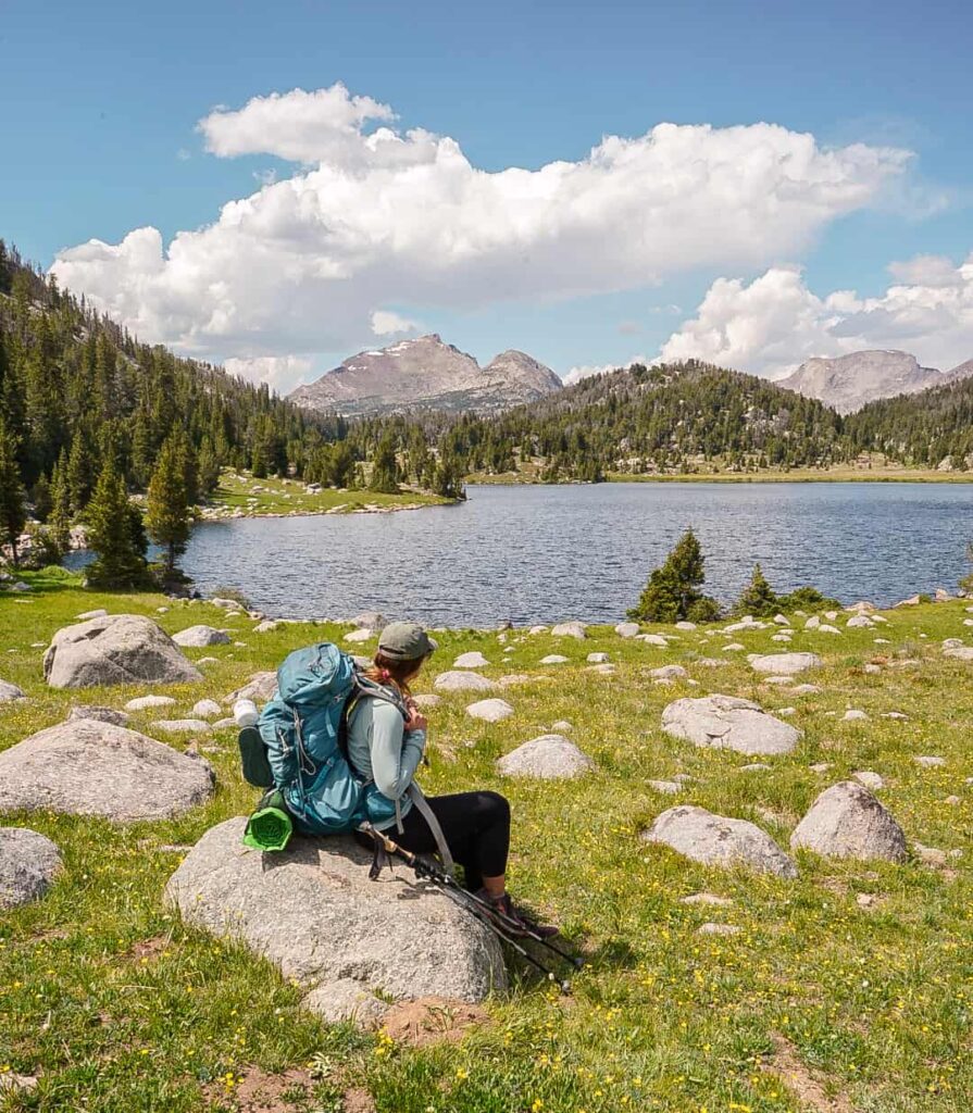 Woman in a light blue sun shirt and black leggings and a large blue backpacking pack sits on a rock with hiking poles leaning against it in front of a lake with mountains in the distance.