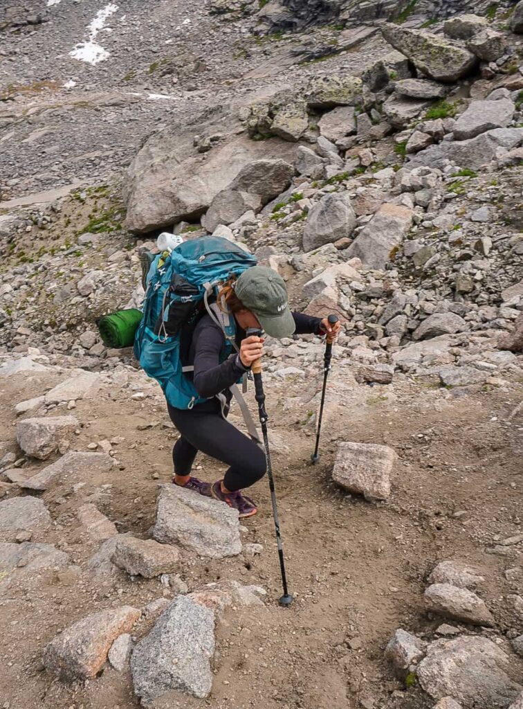 A woman holding hiking poles and wearing a large blue Osprey backpacking pack climbs a steep, rocky hill.