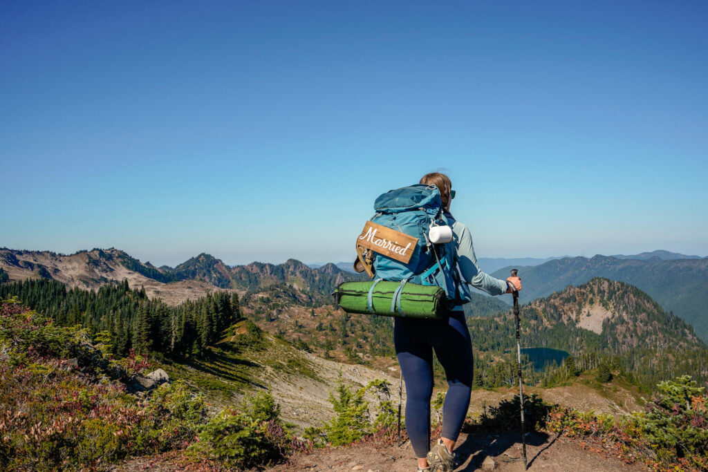 Woman wearing a light blue sun shirt and navy blue leggings and a blue backpacking pack with a married sign on it and a green sleeping pad and mug attached stands facing the Seven Lakes Basin in Olympic National Park while backpacking. She is above the trees and mountains and there is a blue lake down in the distance.