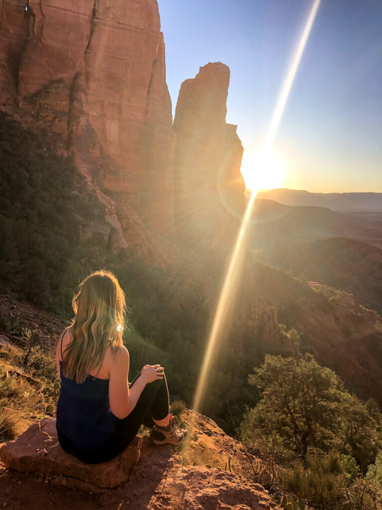 Woman wearing hiking boots sits on a rock overlooking Sedona at sunset with a sun flare bursting from a red rock spire while hiking Cathedral Rock.