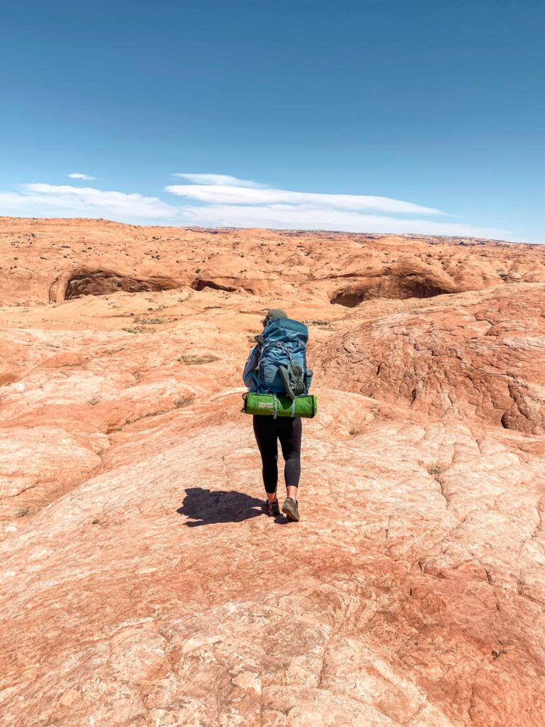 Woman wearing a large blue backpacking pack with a green sleeping pad and Chacos attached walks across salmon colored slickrock towards a canyon in the desert.