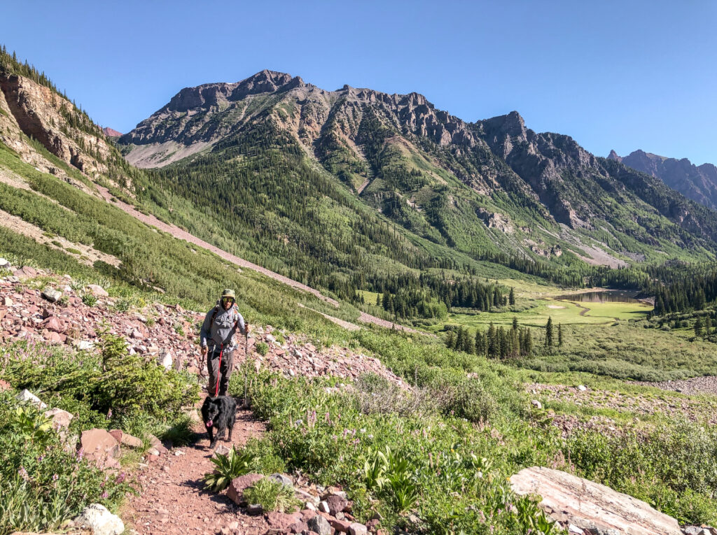 A man backpacking with a black dog on a red leash in front of a verdant valley with mountains behind him in Aspen, Colorado.