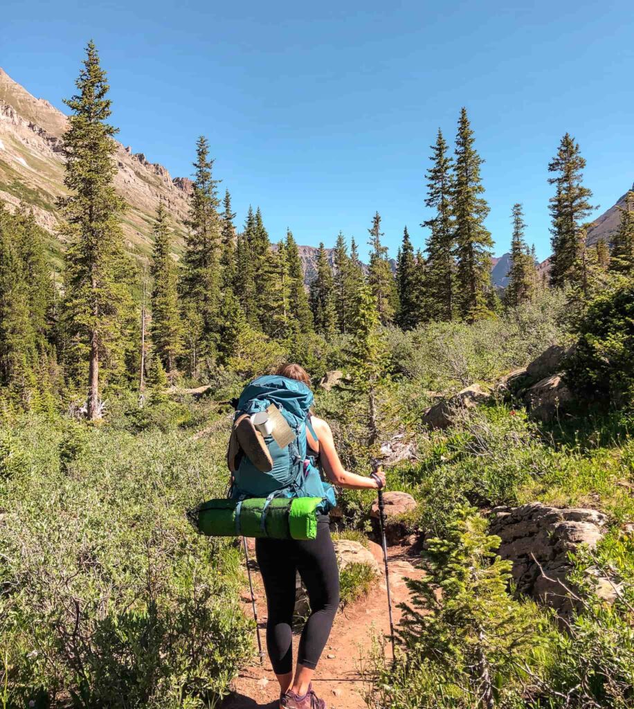 Woman wearing a large blue backpacking pack on an overnight trip in Aspen, Colorado with a green sleeping pad, Birkenstocks, a mug, and permit attached. She holds hiking poles and is standing on a trail with pine trees and mountains around her.