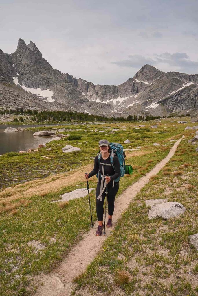 Woman wearing all black with an olive green baseball cap and a large backpacking pack and hiking poles hikes on a trail smiling at the camera with jagged gray, snowcapped mountains behind her while backpacking in the Wind River Range in Wyoming.