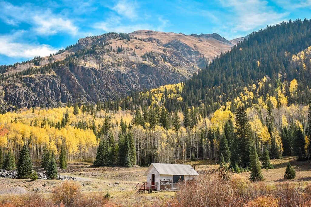 A small white cabin in front of golden aspen trees and evergreens with a mountain peak in the distance along the Million Dollar Highway, which is one of the best scenic drives to see fall colors in Colorado.