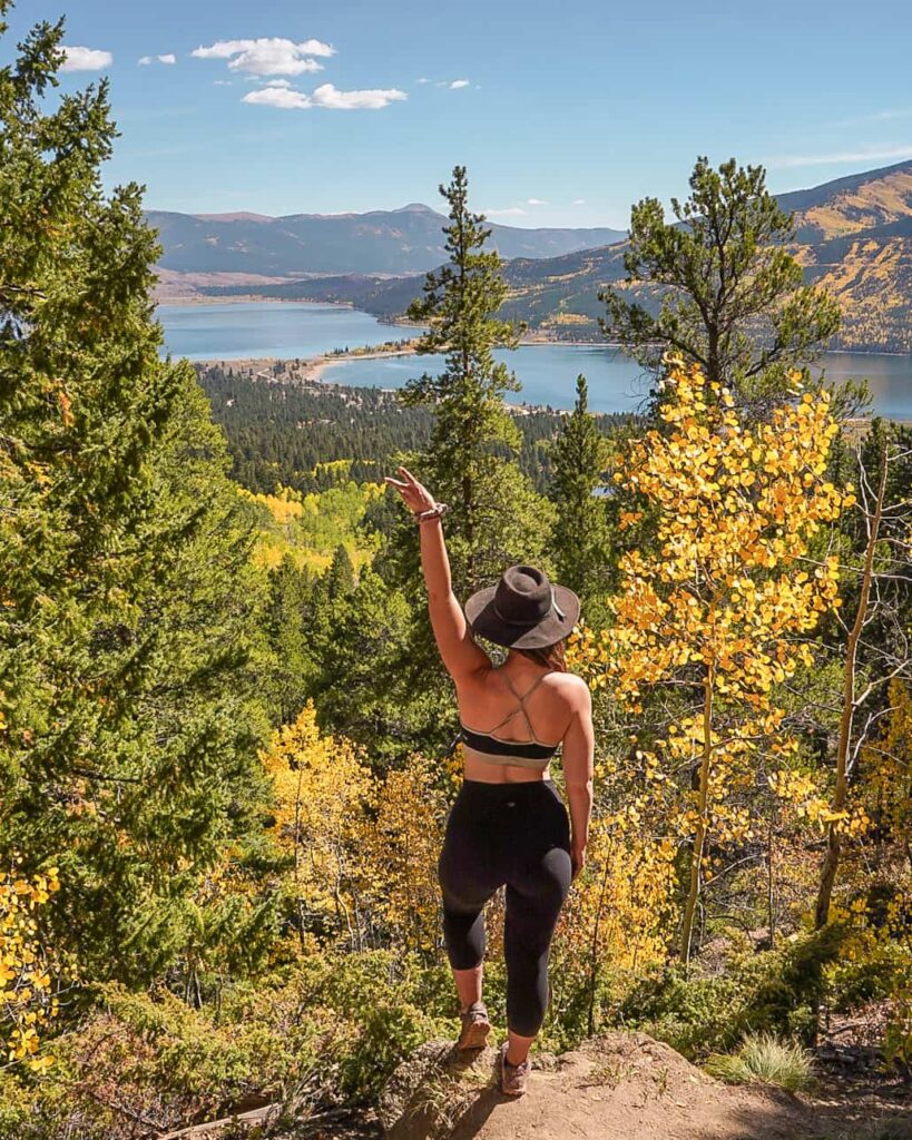 Woman stands with a peace sign in the air overlooking twin lakes and aspen trees in the fall in Colorado.