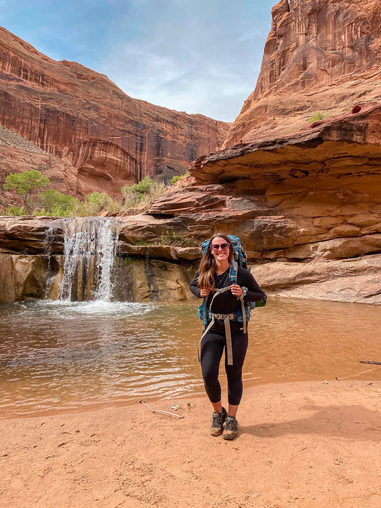 Woman wearing a long sleeve black shirt, black leggings, sunglasses, and a large blue backpacking pack stands in front of a waterfall in an orange sandstone canyon while backpacking in Coyote Gulch in Escalante, Utah.