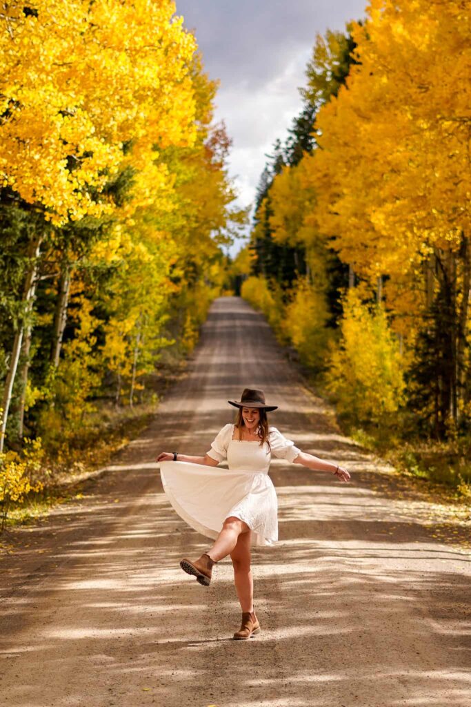 Woman in a white dress takes photos in the road during fall in Colorado surrounded by golden aspen trees.