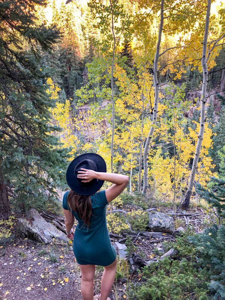 Woman in a green dress and black hat stands in front of aspen trees while on one of the best scenic drives for fall colors in Colorado.