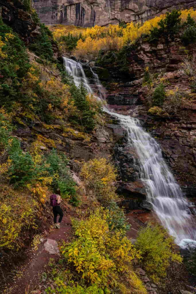 Woman wearing a maroon backpack and leggings stands on a trail to the left of a waterfall in a canyon in Telluride in the fall.