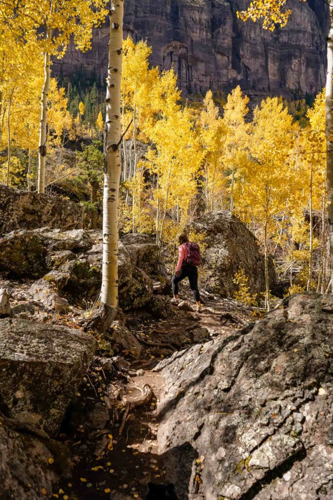 A woman in a pink top climbs a rocky trail surrounded by golden aspen trees.